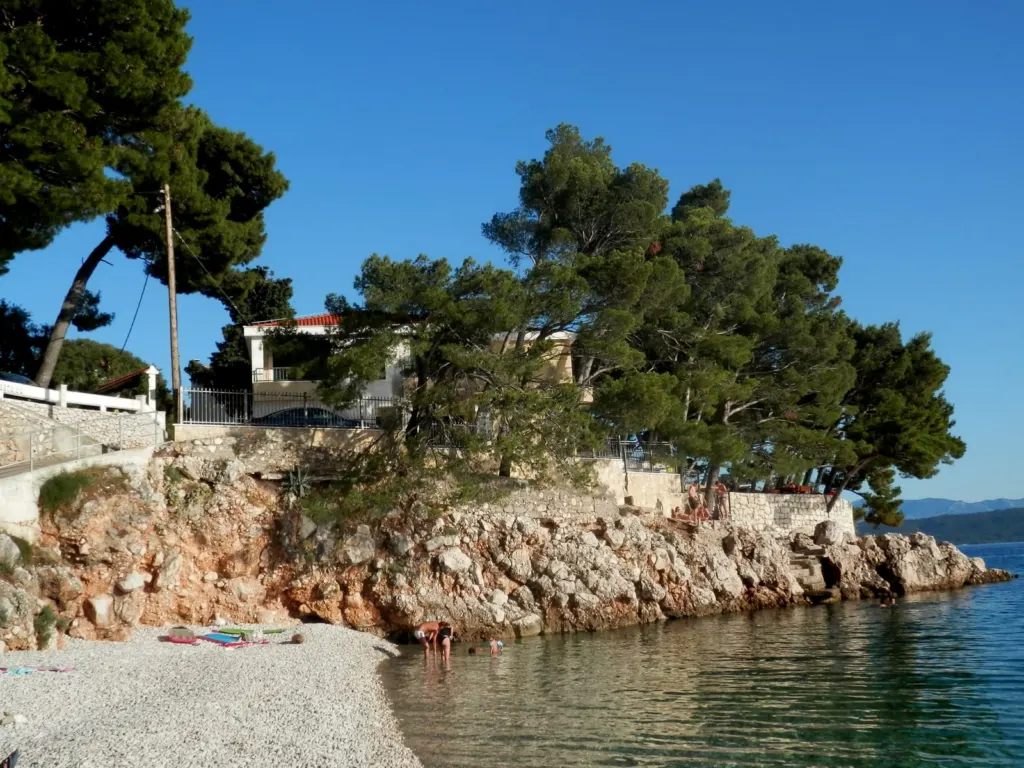 Šljunčana plaža u Portu, s kupačima u plitkom moru, stjenovitim rtom i borovima iznad mora